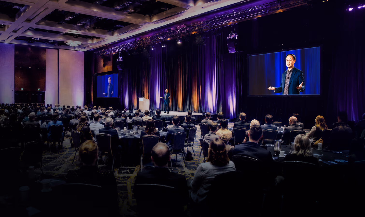 Speaker presenting on stage to a large audience seated at round tables in a conference hall.