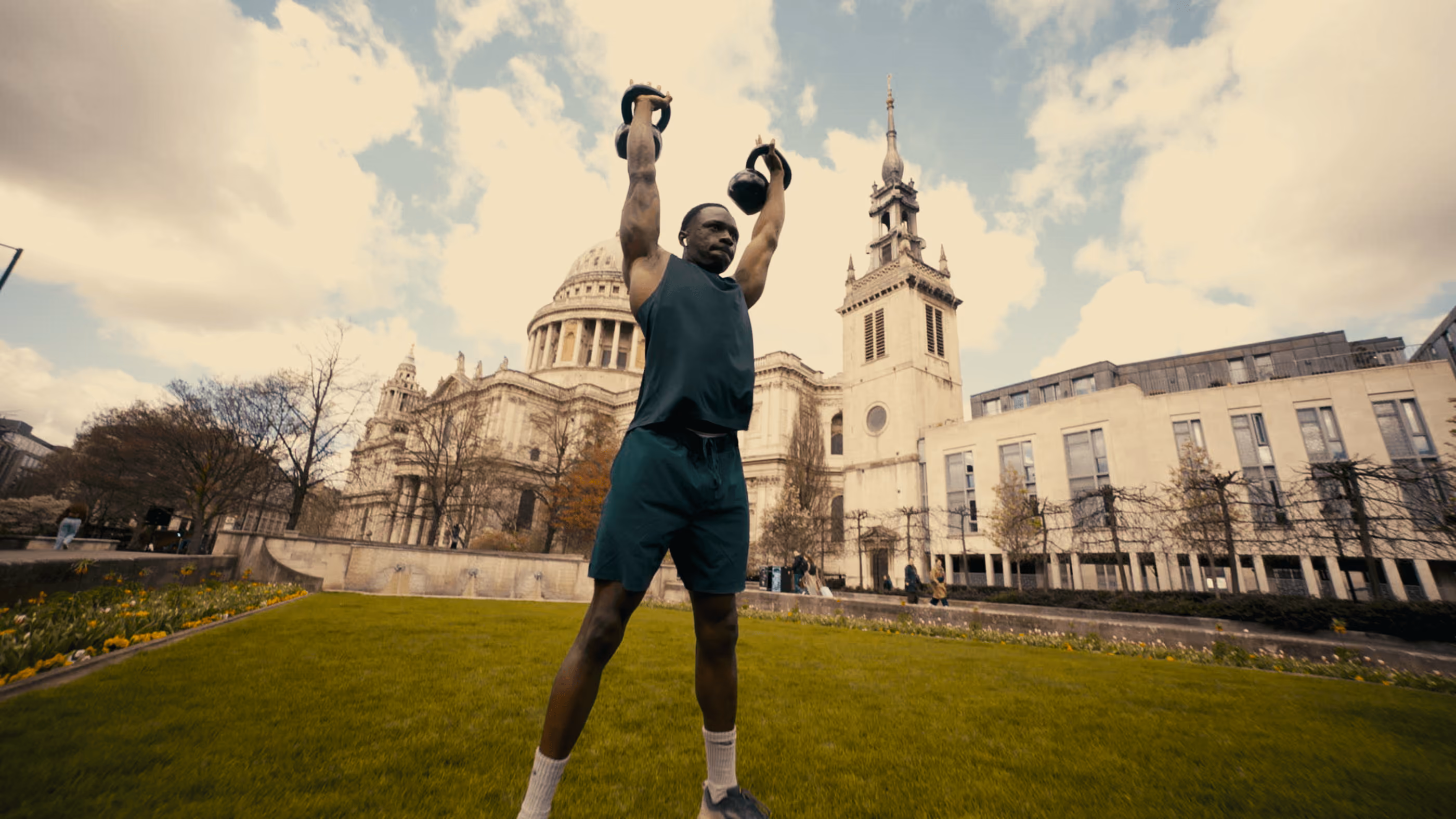 Man in athletic wear lifts two kettlebells overhead on green lawn near historic cathedral.
