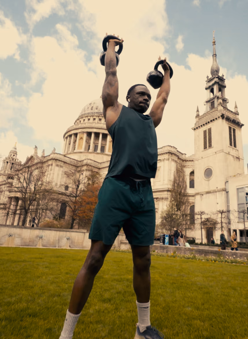 Man exercising outdoors lifting two kettlebells overhead with historic buildings in the background.