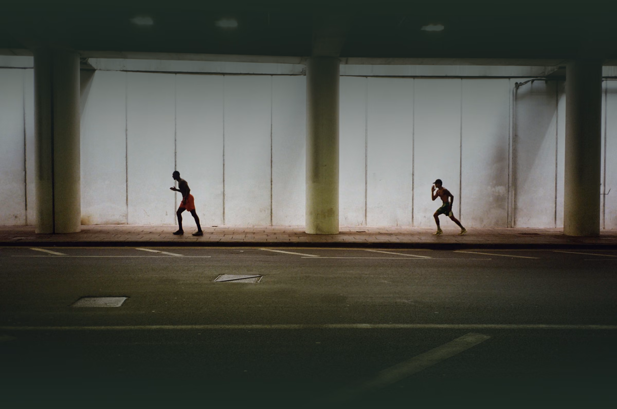 Two men running in an underpass with pillars and a white wall in the background.