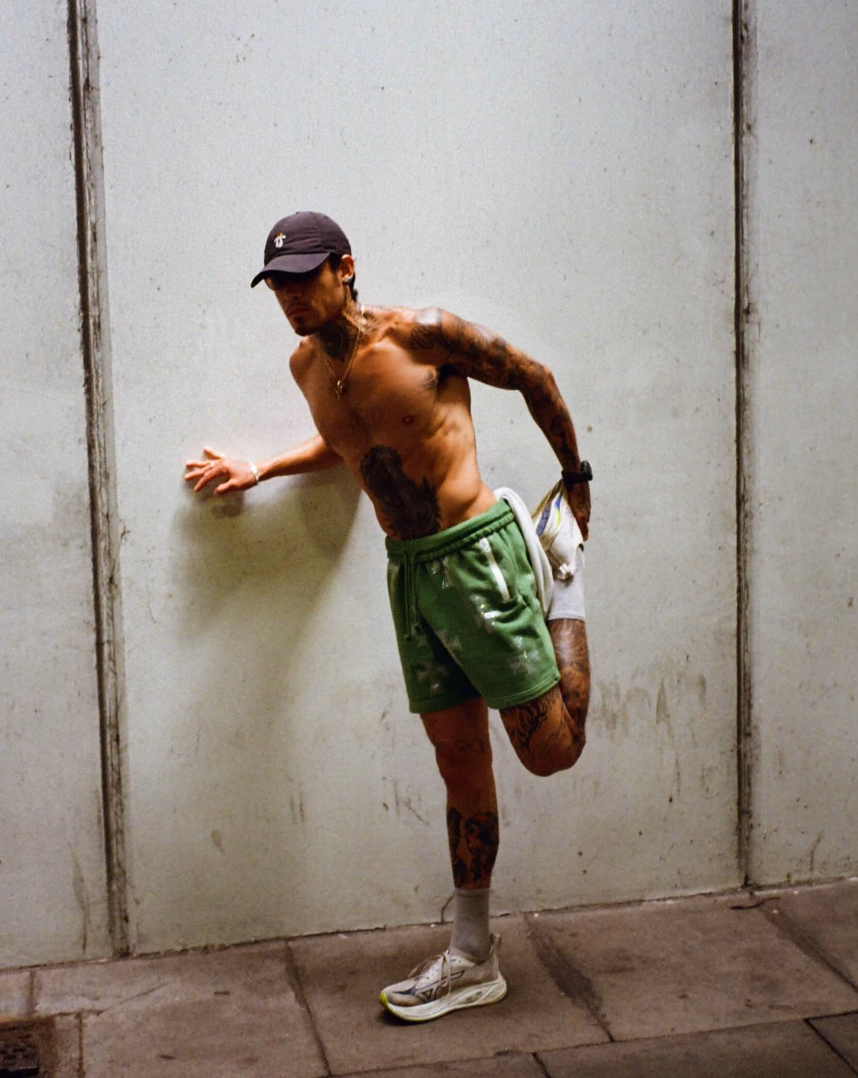 Shirtless man wearing green shorts and cap stretches his leg against a wall indoors.