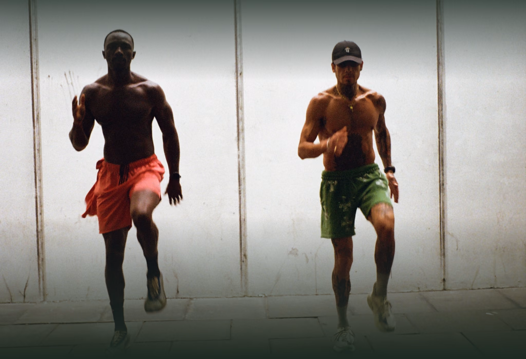 Two shirtless men running in place against a white wall, wearing red and green shorts.