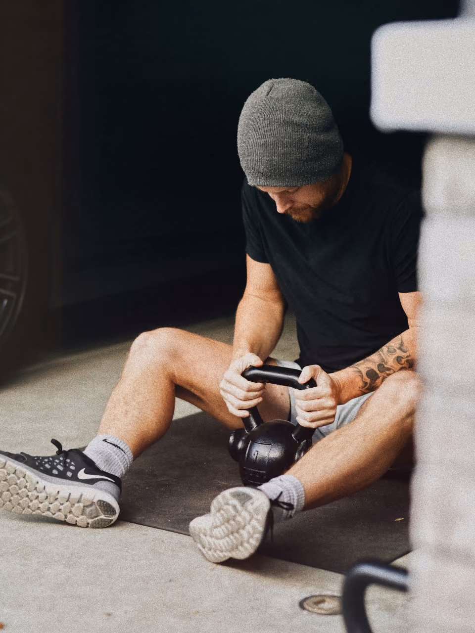 Man in a gray beanie and black shirt sitting on floor holding a kettlebell with tattooed arm visible.