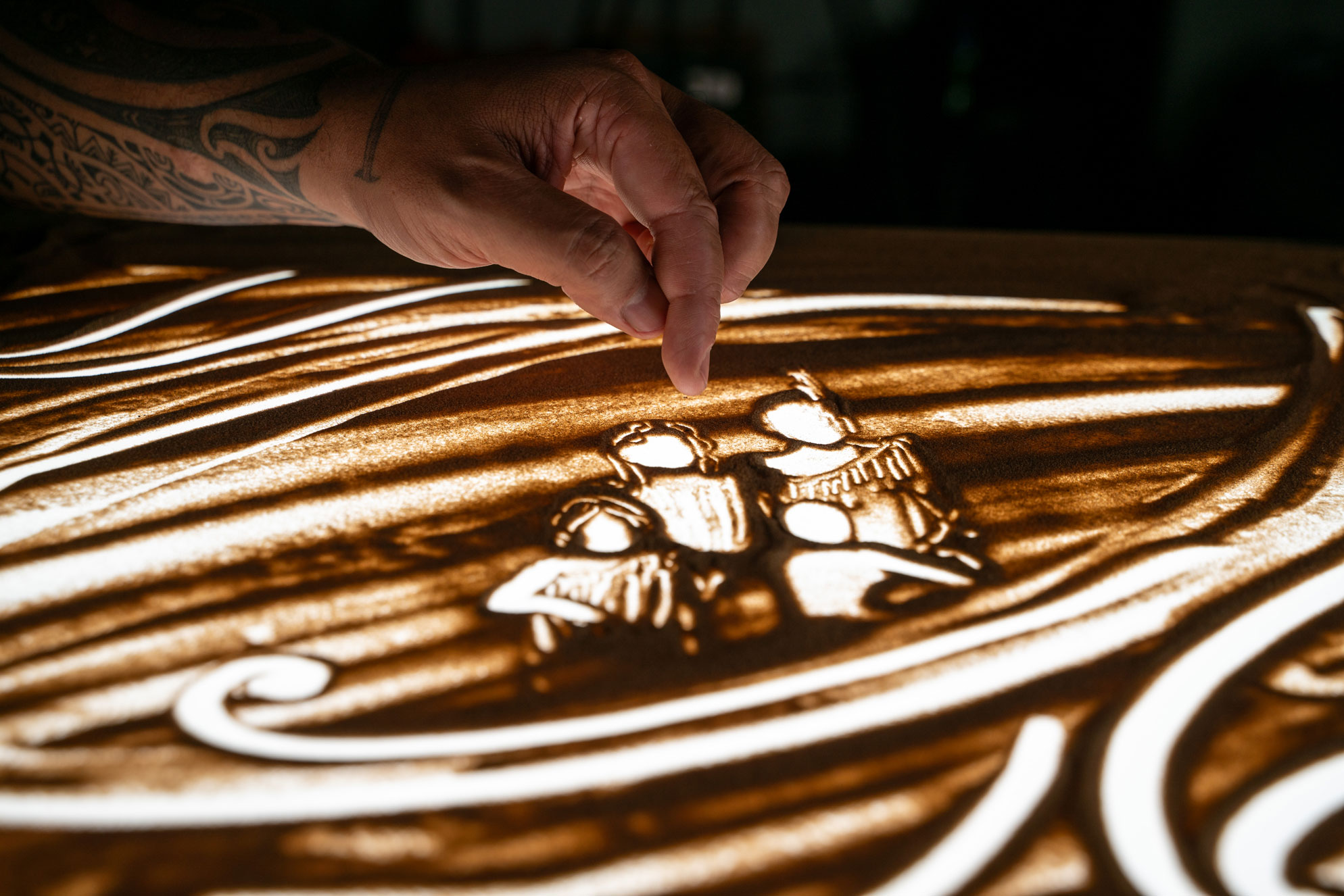 Hand with tattoo creating figures of four people in sand art with light and shadow effects.