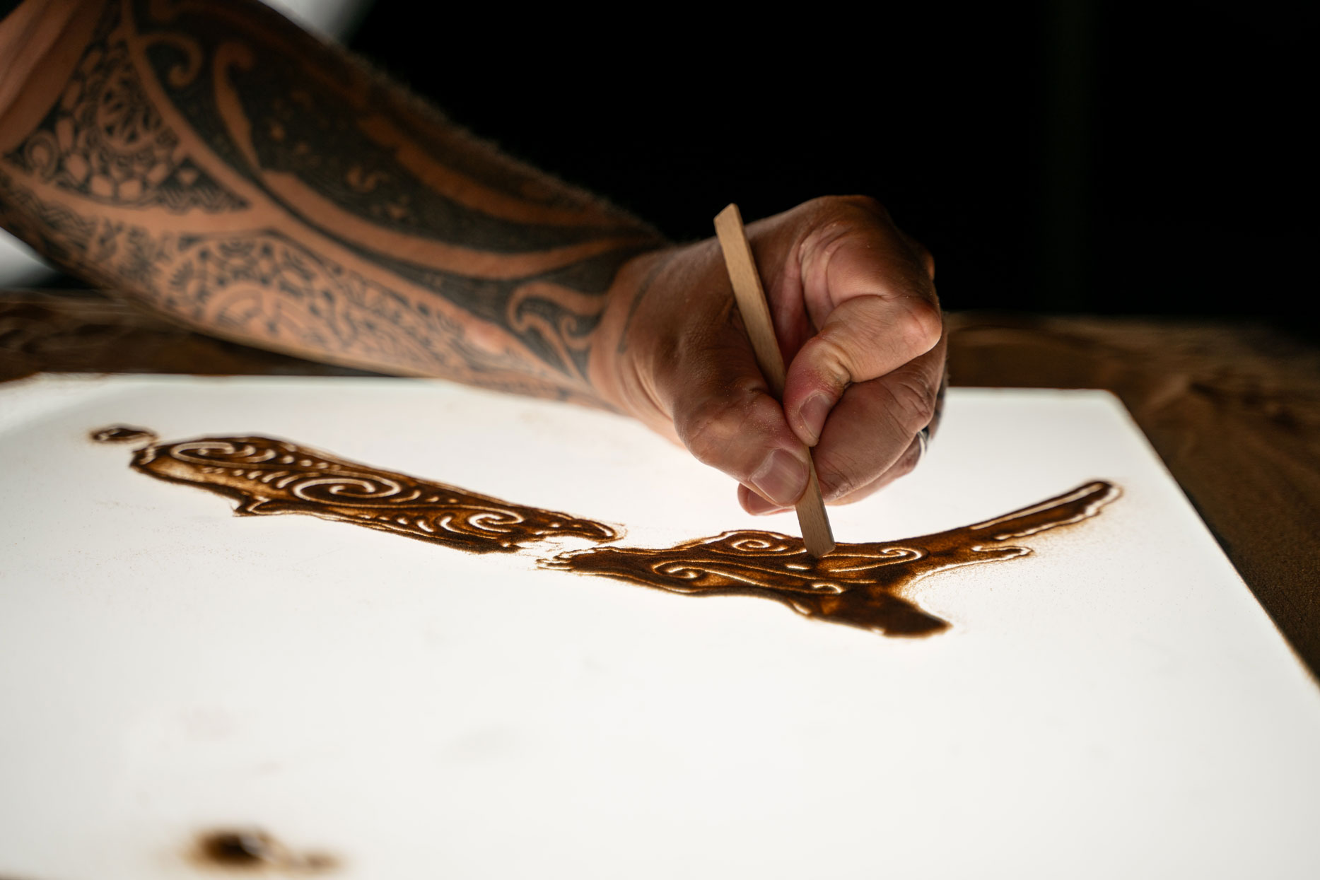 Hand with tattooed arm carving Maori-style patterns on sand with a wooden stick.
