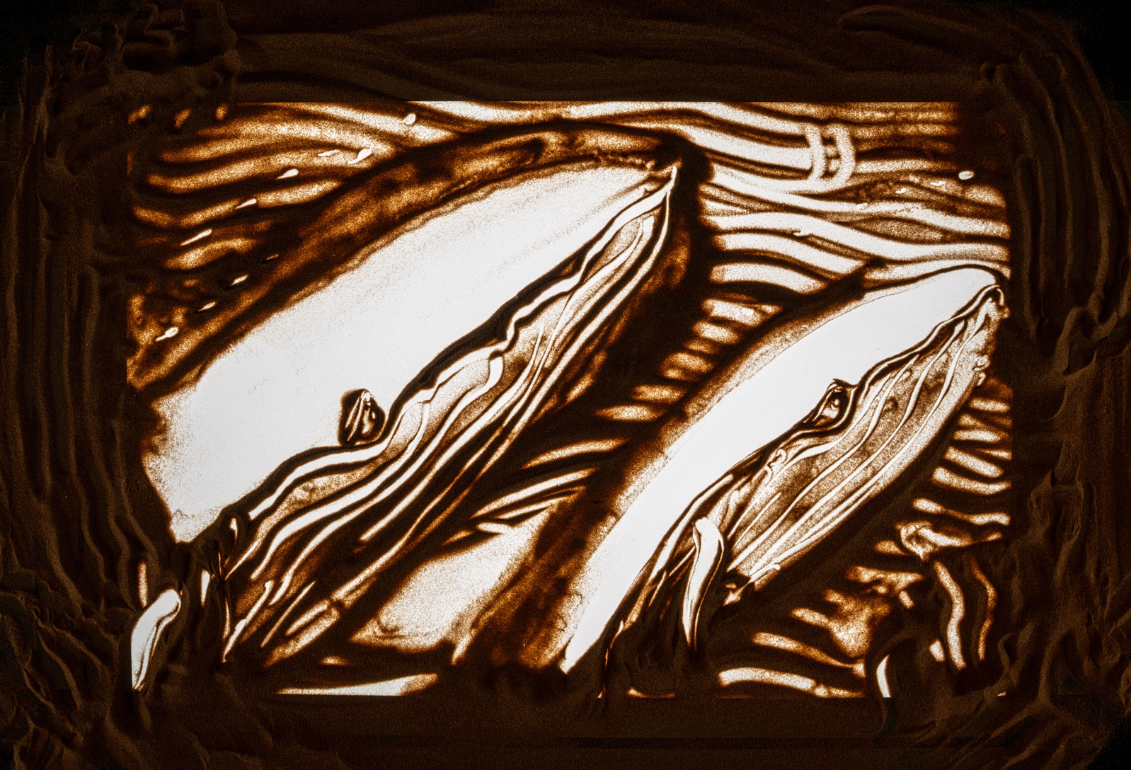 Sand art of two whales swimming side by side with flowing lines representing water.