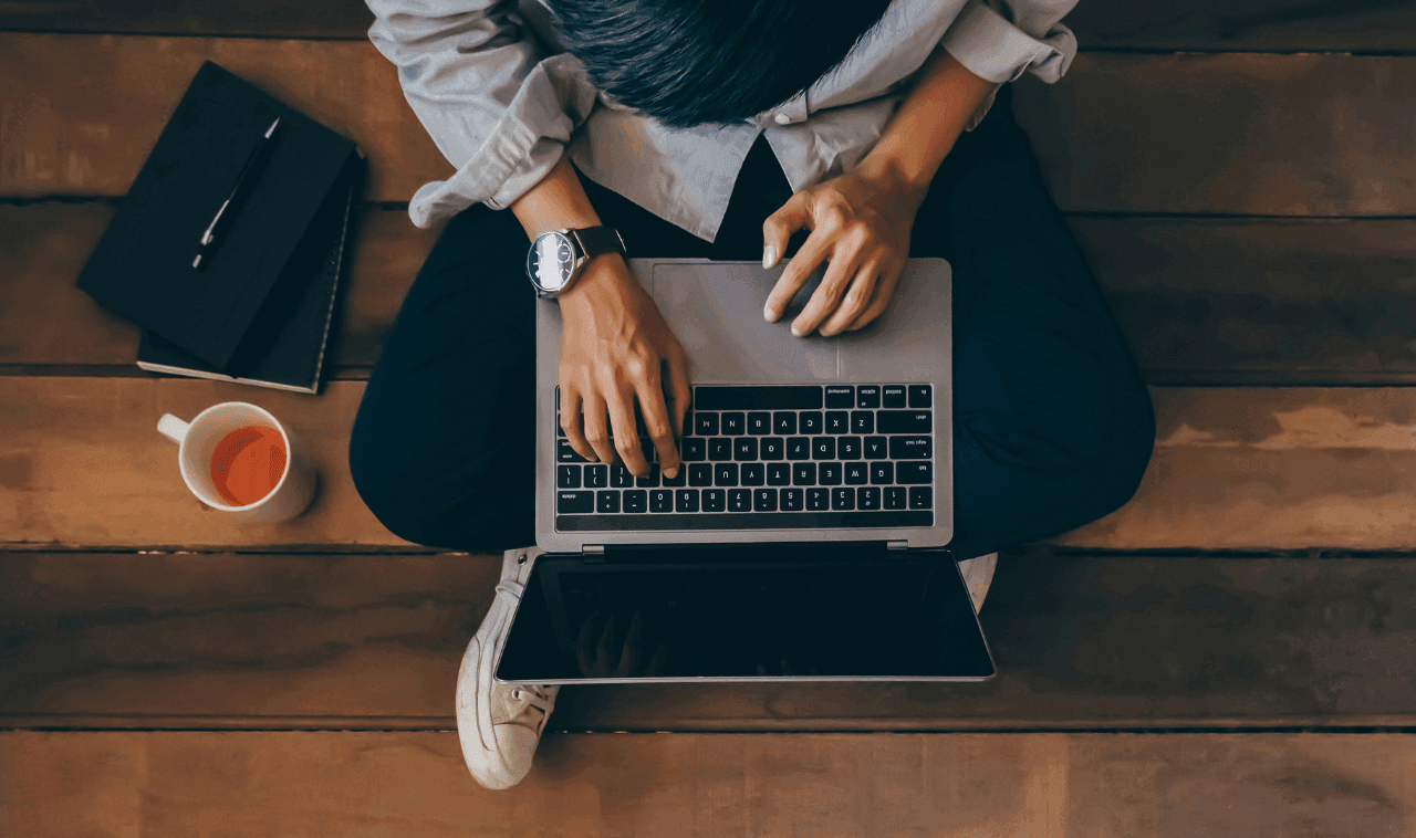 Man sitting on a wooden floor on his computer with a cup of tea and a notebook sitting on the floor next to him.