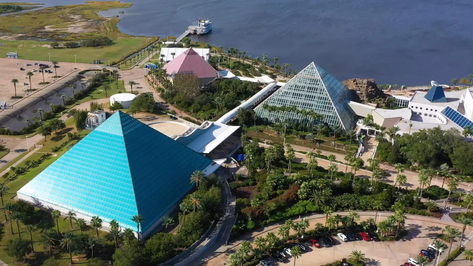 Aerial view of glass pyramids surrounded by greenery beside a large water body with a docked boat.