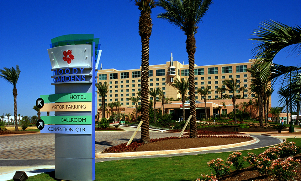 The Moody Gardens Hotel exterior and entrance signpost, framed by palm trees.