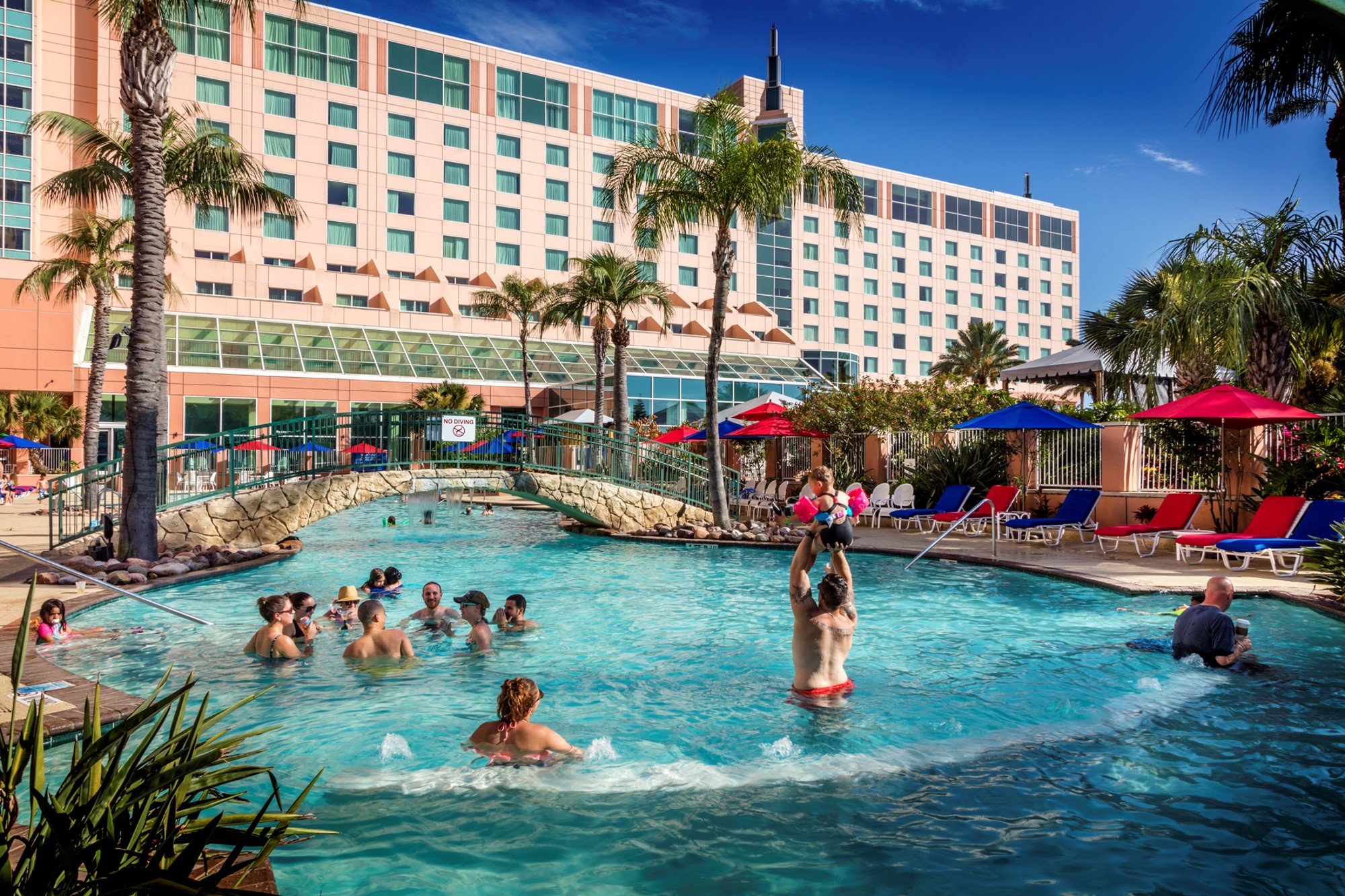 Guests relaxing in the Moody Gardens Hotel resort pool with red and blue umbrellas.