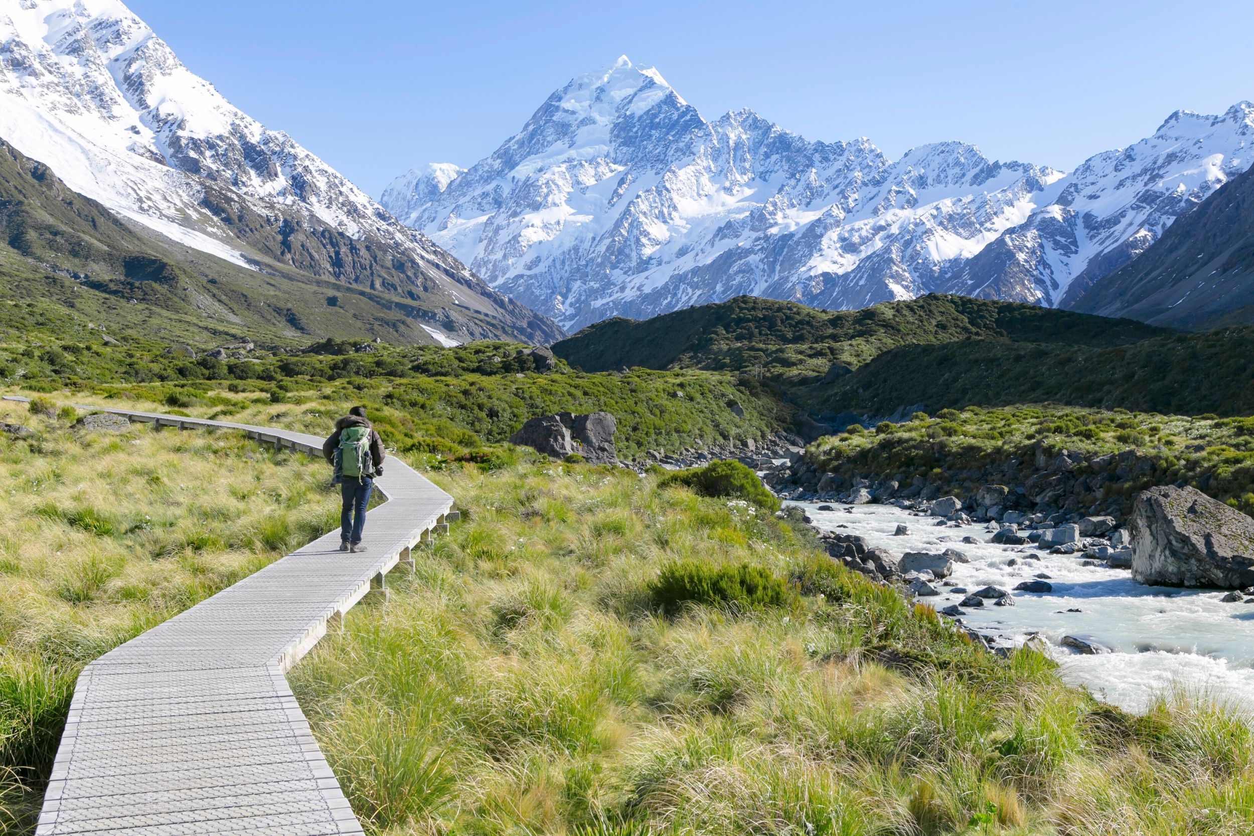 Person hiking on boardwalk trail beside stream with snowy mountain in background under blue sky.