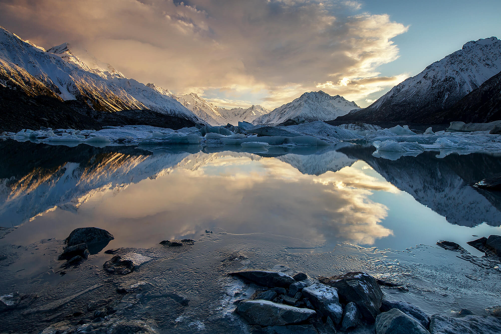 Snowy mountains reflected in a calm lake with floating icebergs under a cloudy sky at sunset.