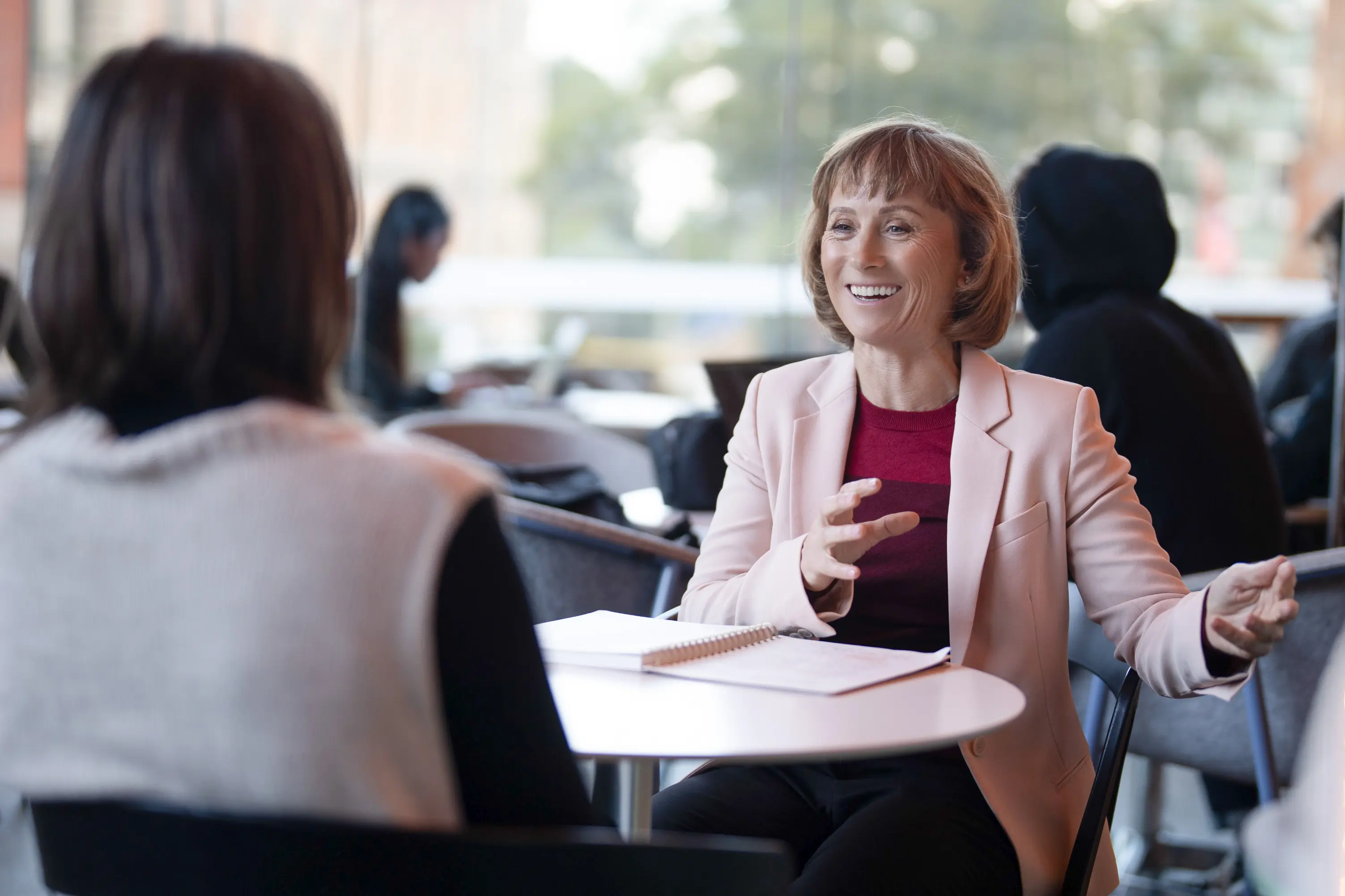Smiling woman in a pink blazer talking with another person at a cafe table with a notebook.