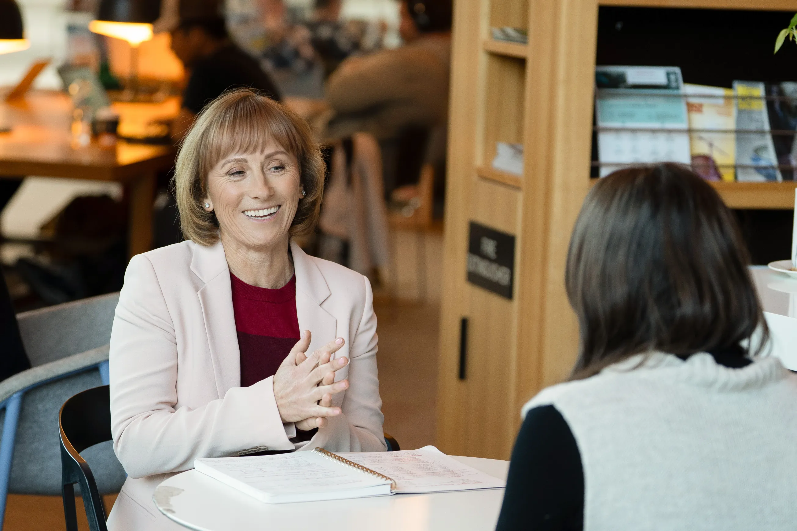 Smiling woman in beige blazer talking to another person at a table with an open notebook.