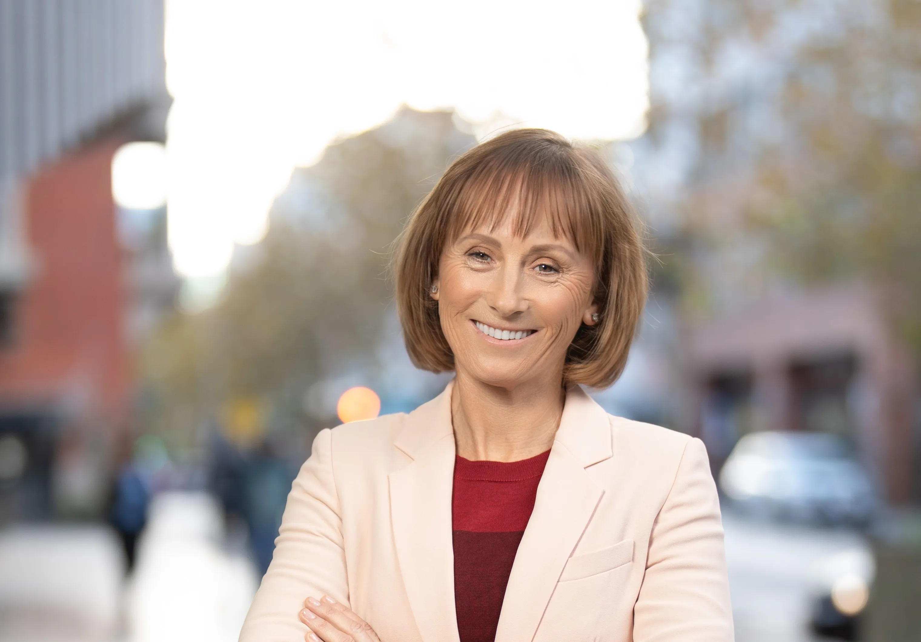 Smiling middle-aged woman in pale blazer with arms crossed standing on a bright city street.