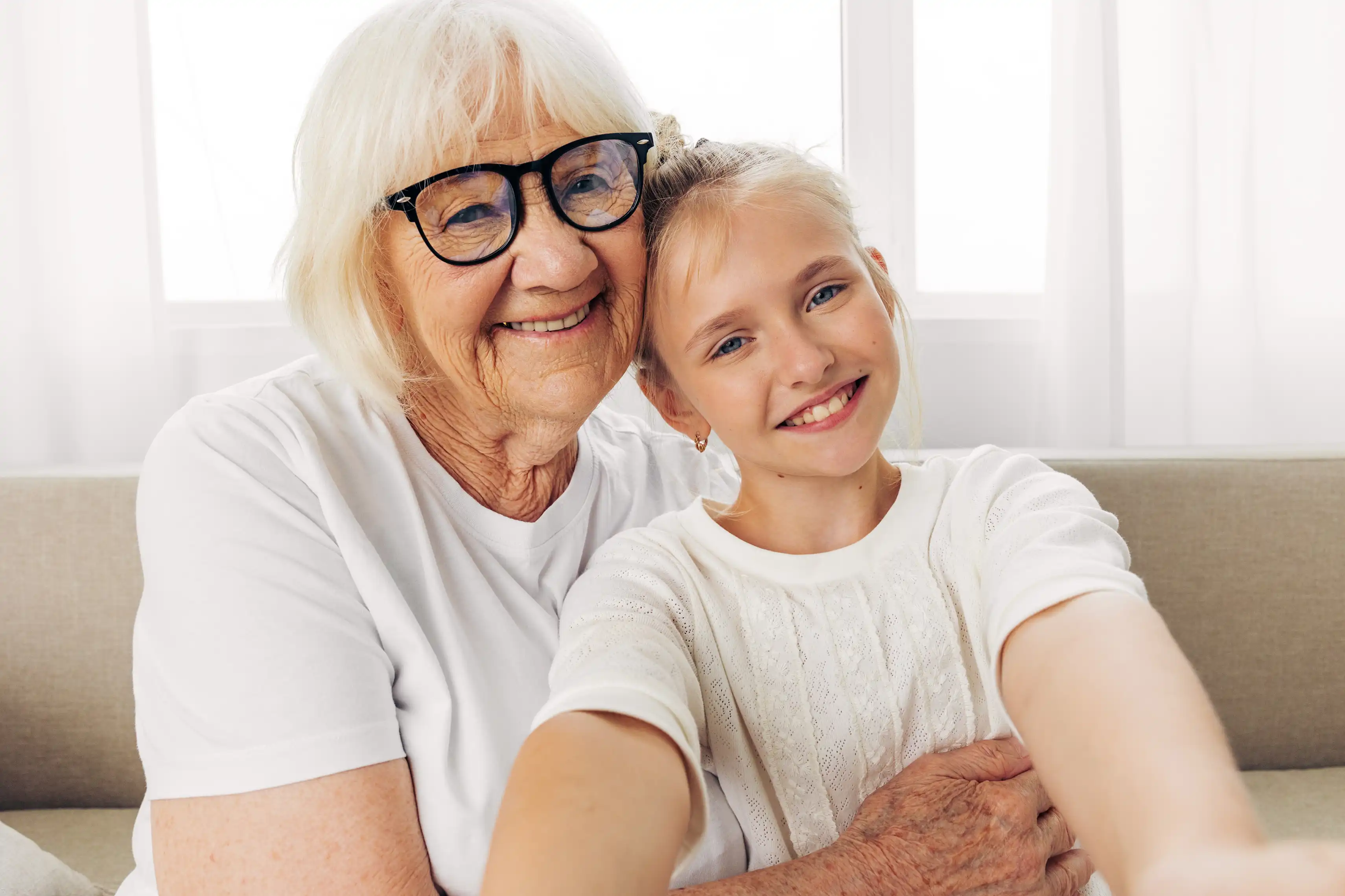 Elderly lady with glasses smiling while hugging her grand daughter on a couch
