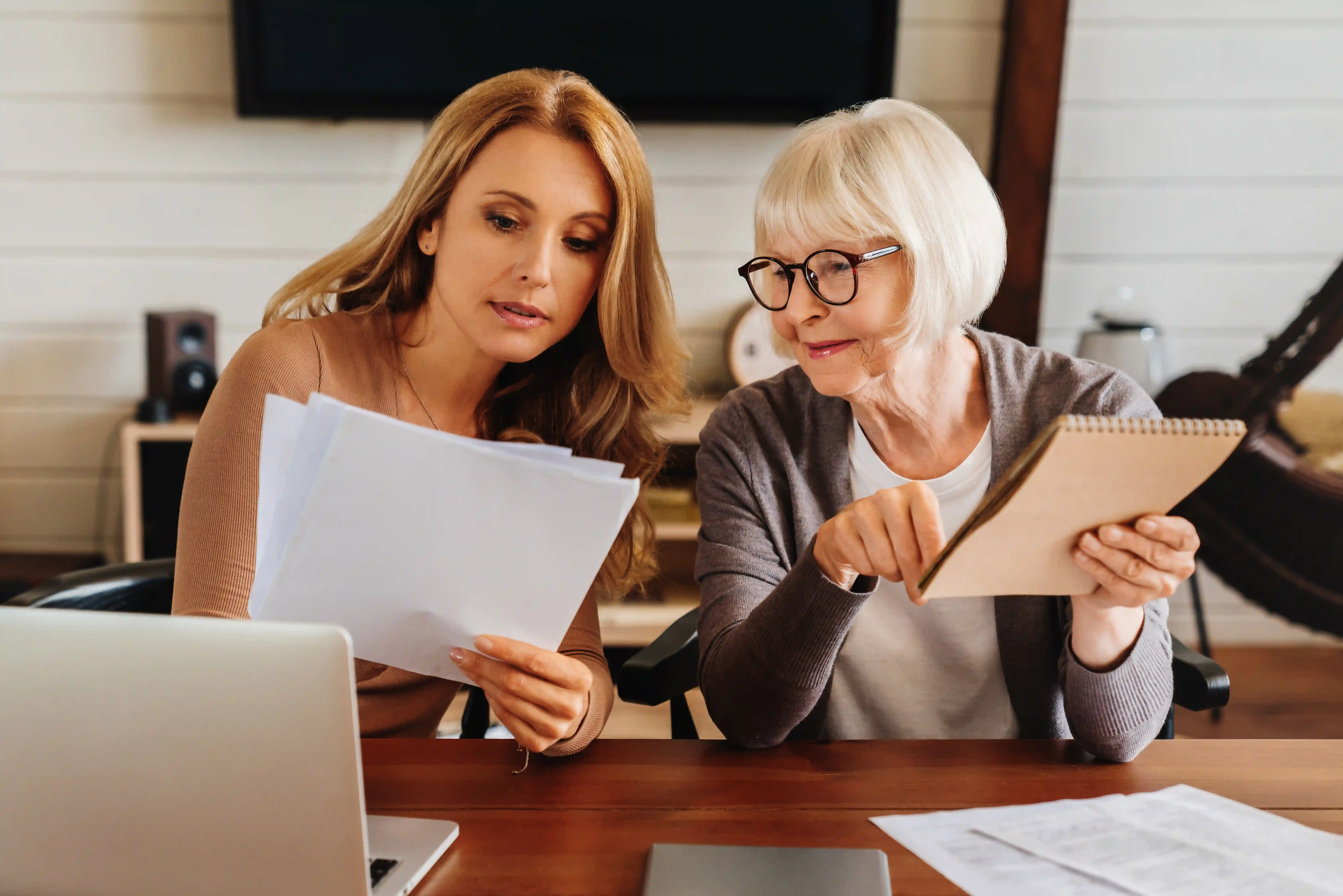 Mature daughter helping her mother with paperwork on a kitchen table with a laptop