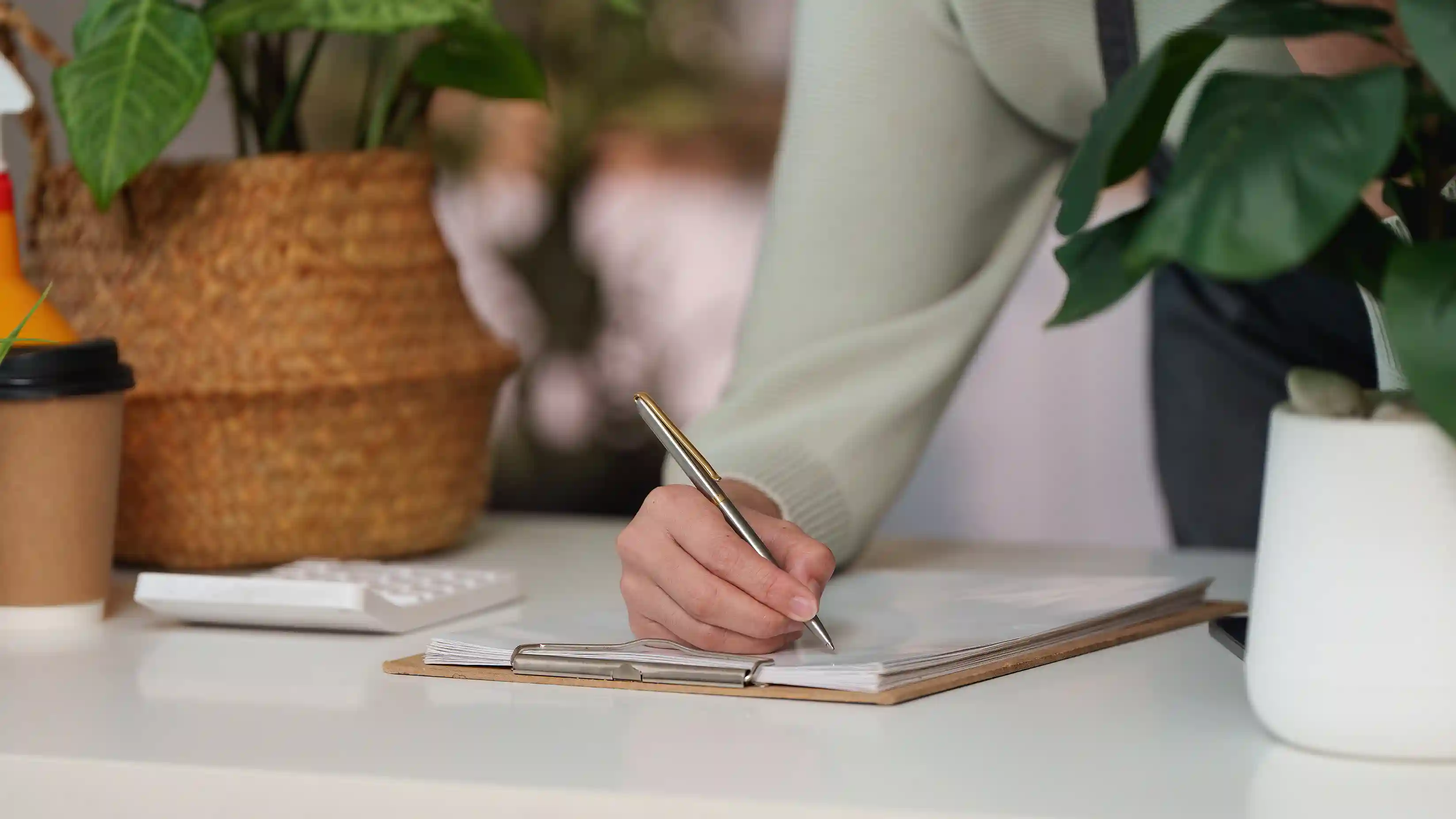 Woman signing paperwork on a clipboard with a coffee and plant on the desk beside her