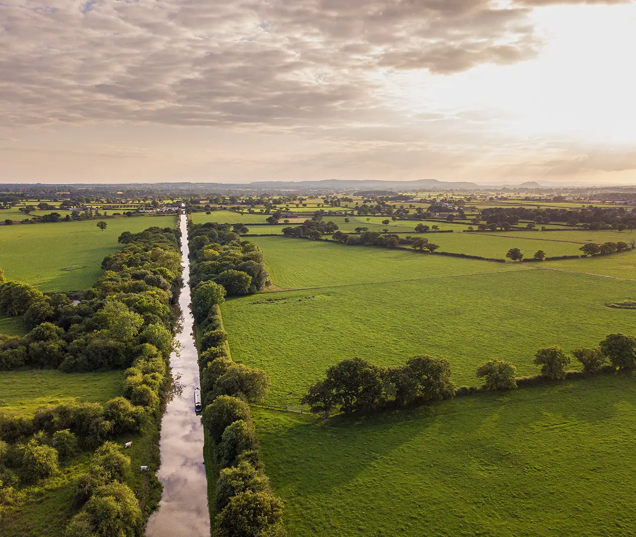 Aerial view of a canal with a boat surrounded by green fields and trees under a cloudy sky.