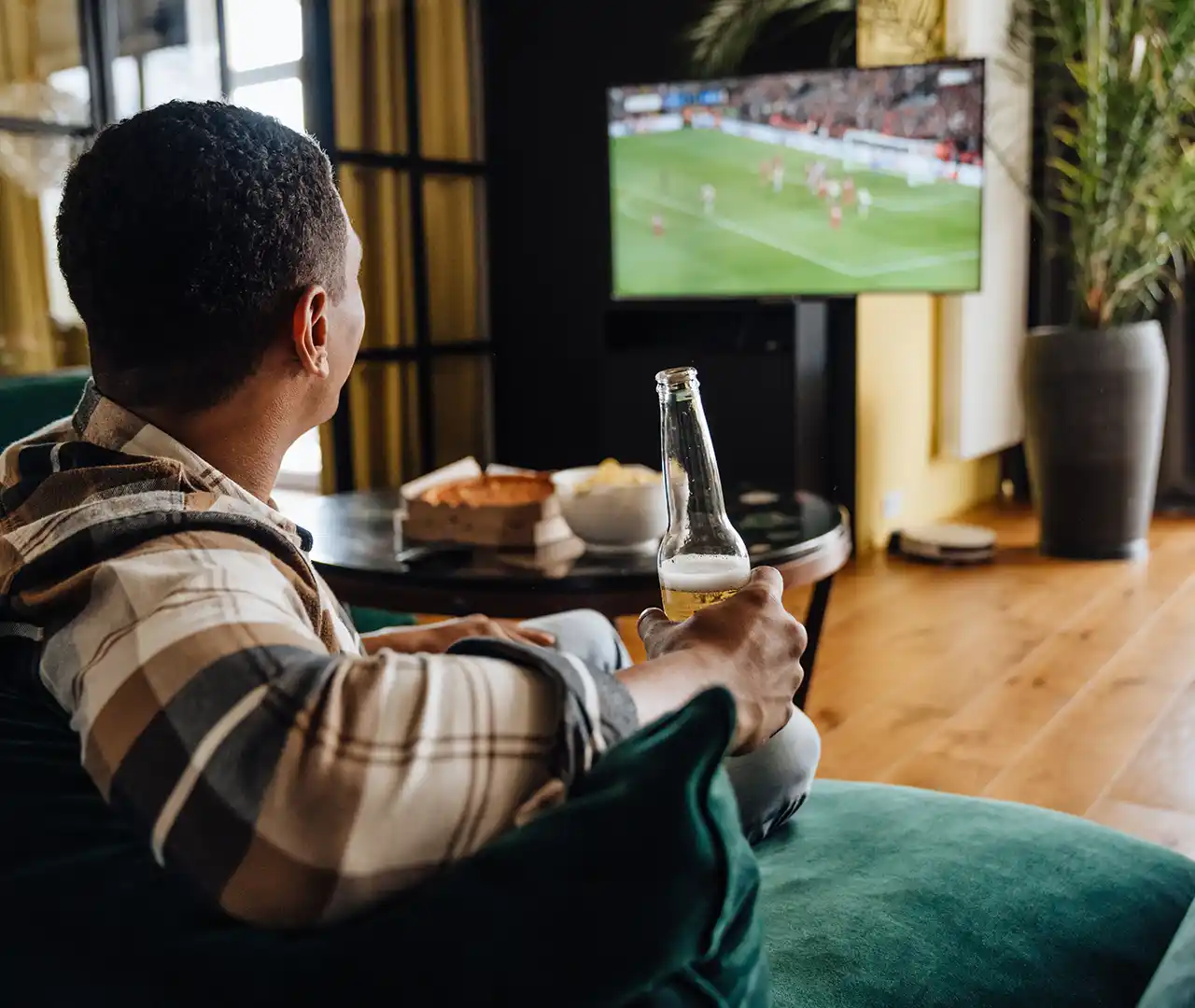 Man sitting on couch holding beer bottle watching football on TV with snacks on table.