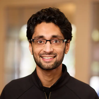 Smiling man with glasses and curly dark hair wearing a black zip-up jacket indoors.