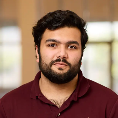Portrait of a man with dark hair and beard, wearing a maroon collared shirt, with a softly blurred background.