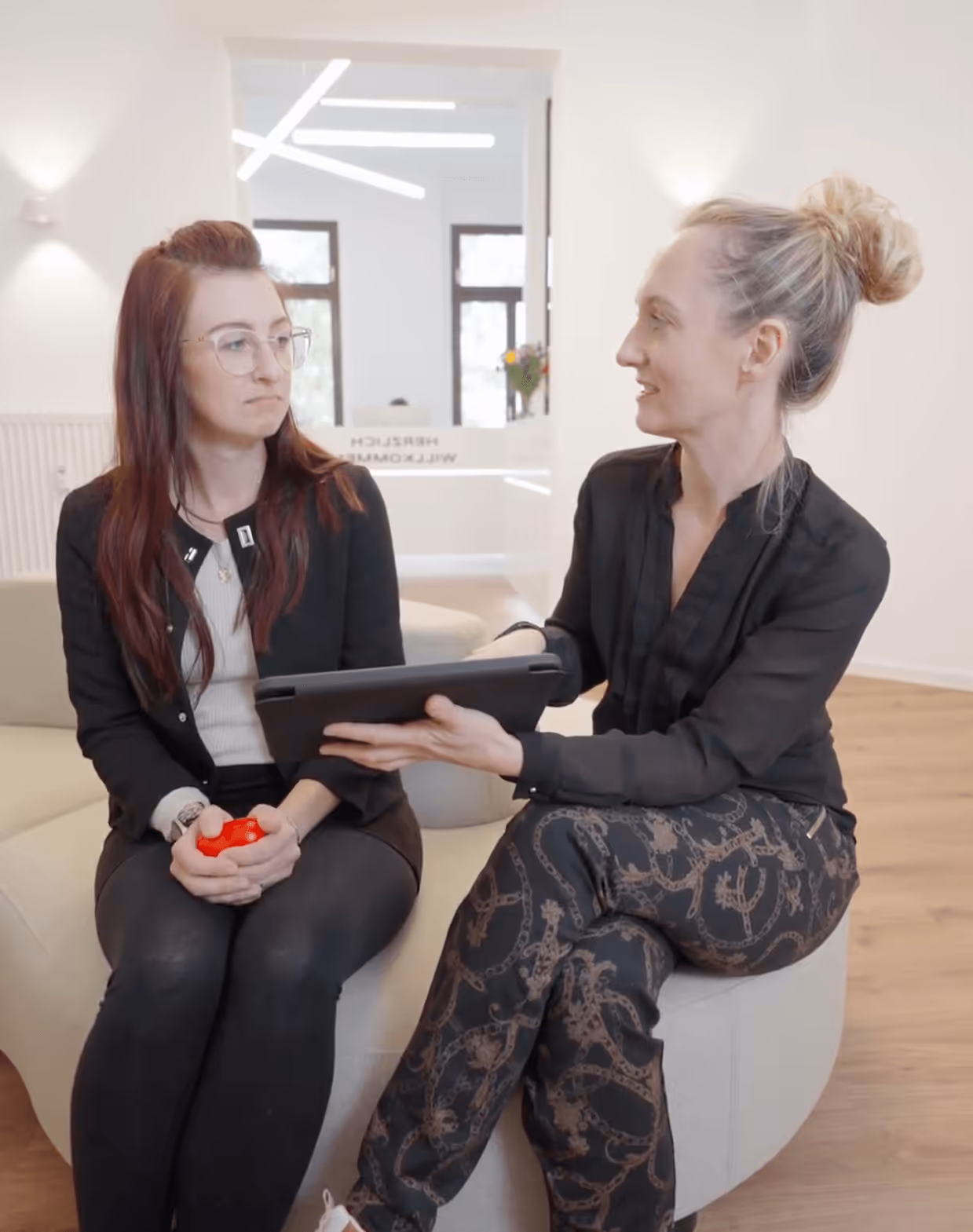 Zwei Frauen im Gespräch auf einem Sofa in einem modernen, hellen Raum; eine hält ein Klemmbrett, die andere einen Stressball.