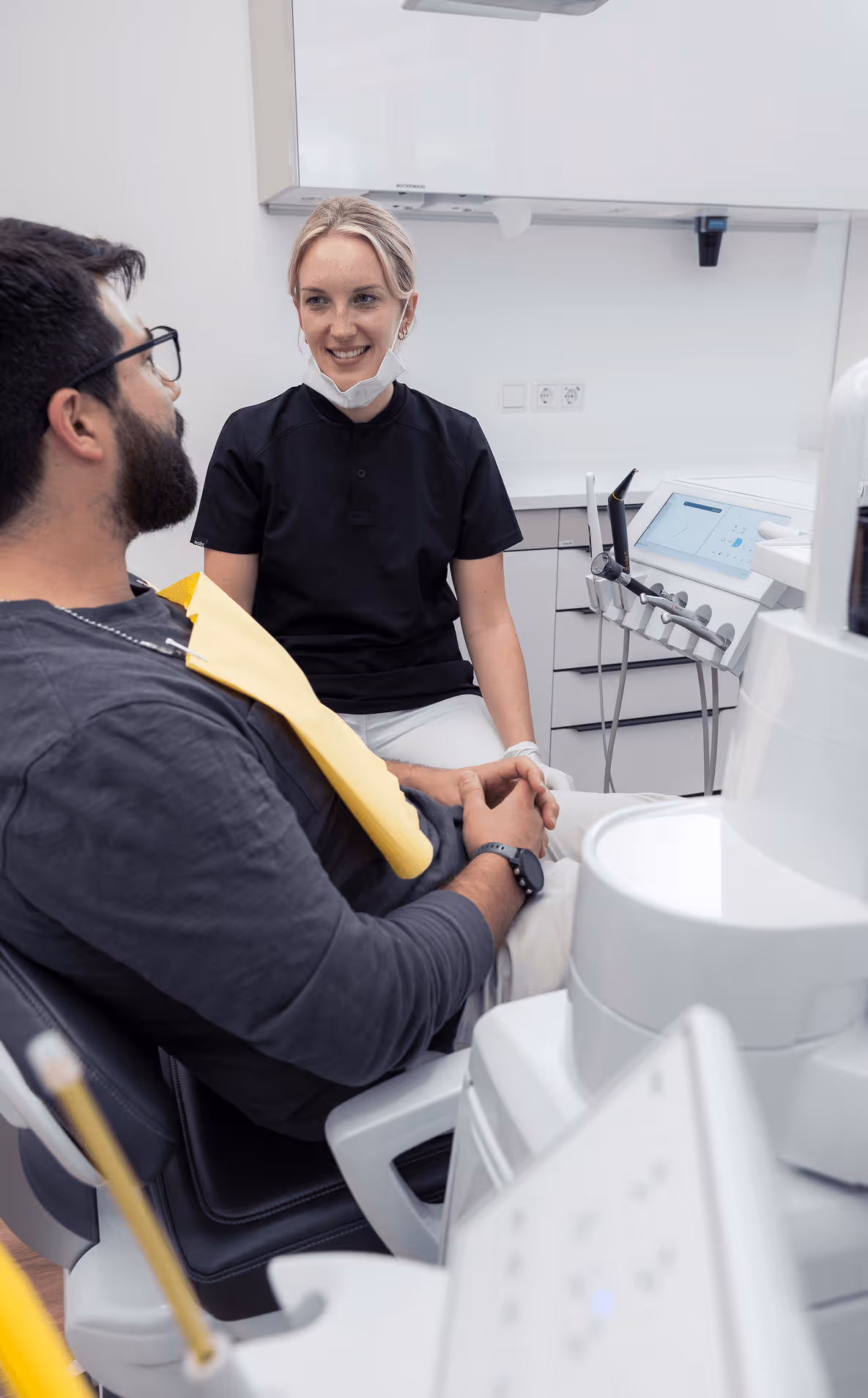 Zahnärztin spricht mit männlichem Patienten im modernen Behandlungszimmer. Beide lächeln, entspannte Atmosphäre.
