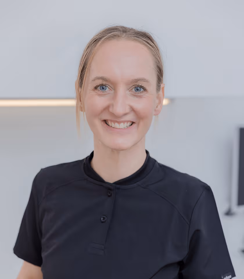 Frau in schwarzem Polohemd lächelt freundlich in neutralem Innenraum.