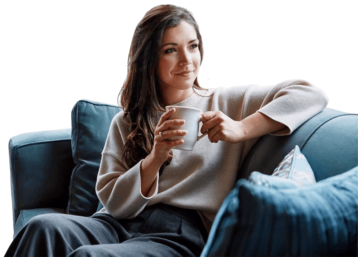 Smiling woman with long brown hair sitting on a blue couch, holding a white mug with both hands, looking thoughtfully to the side.