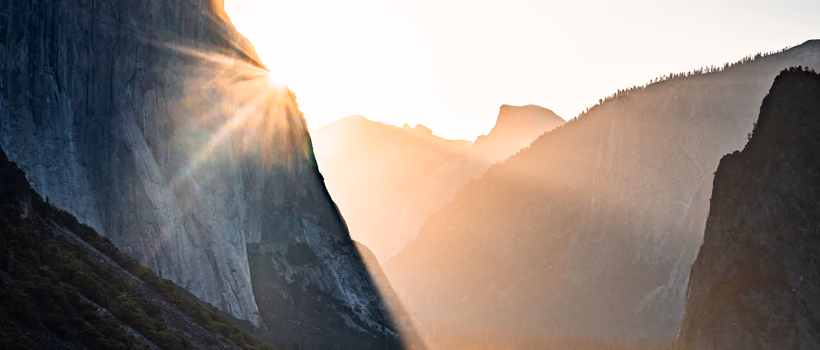 Sunlight shining over a steep mountain cliff with a hazy mountain range in the background during sunrise or sunset.