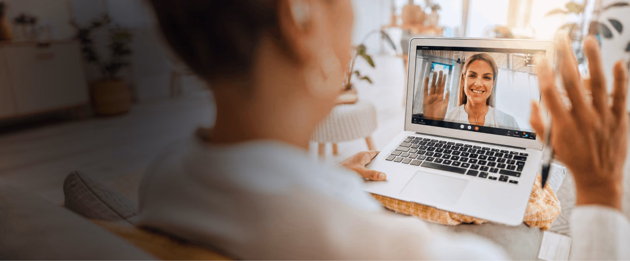 Person sitting indoors holding a laptop showing a video call with a smiling woman waving.
