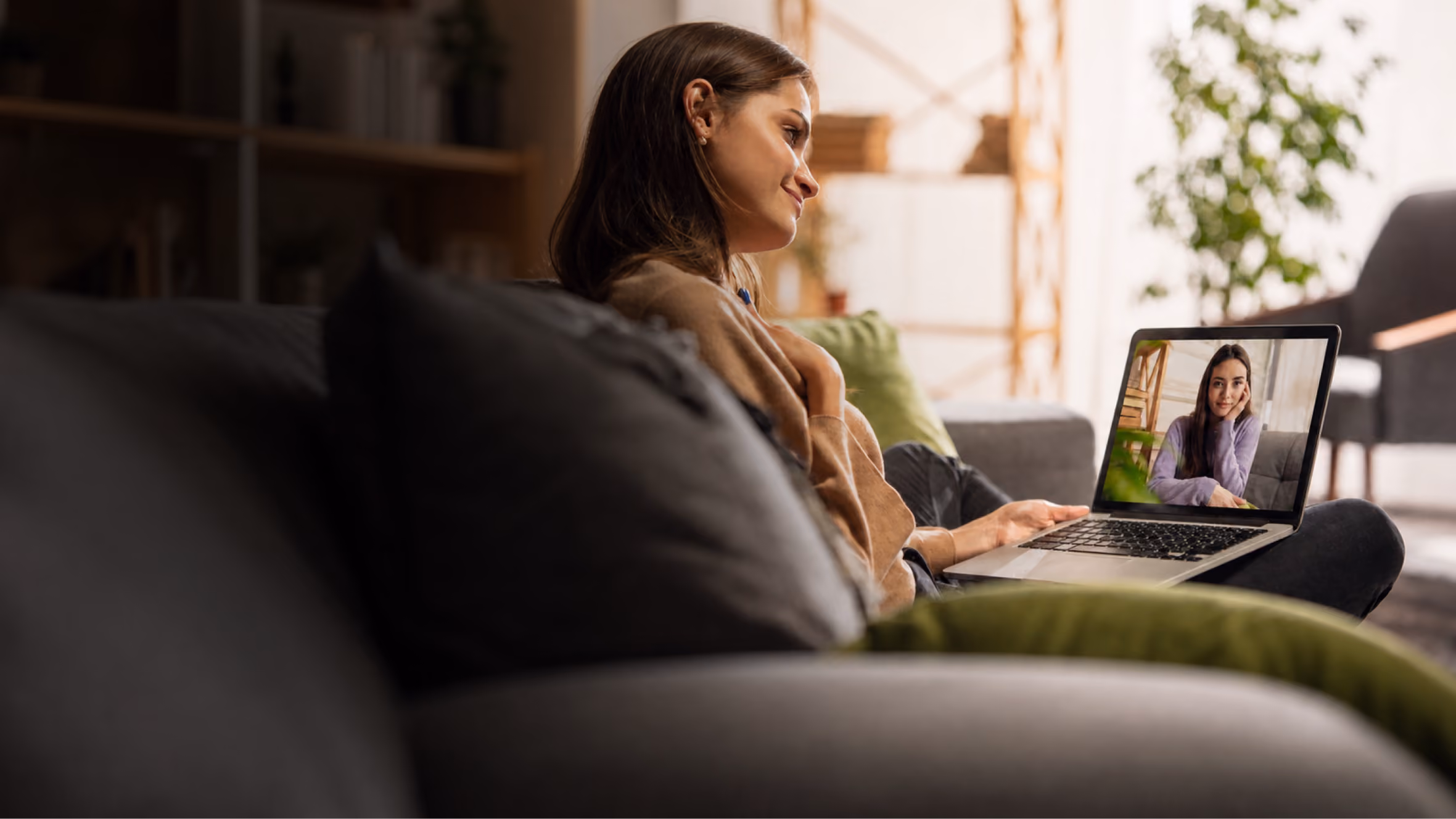 A woman sitting on a couch holding a laptop on her lap, having a video call with another woman who is visible on the laptop screen.