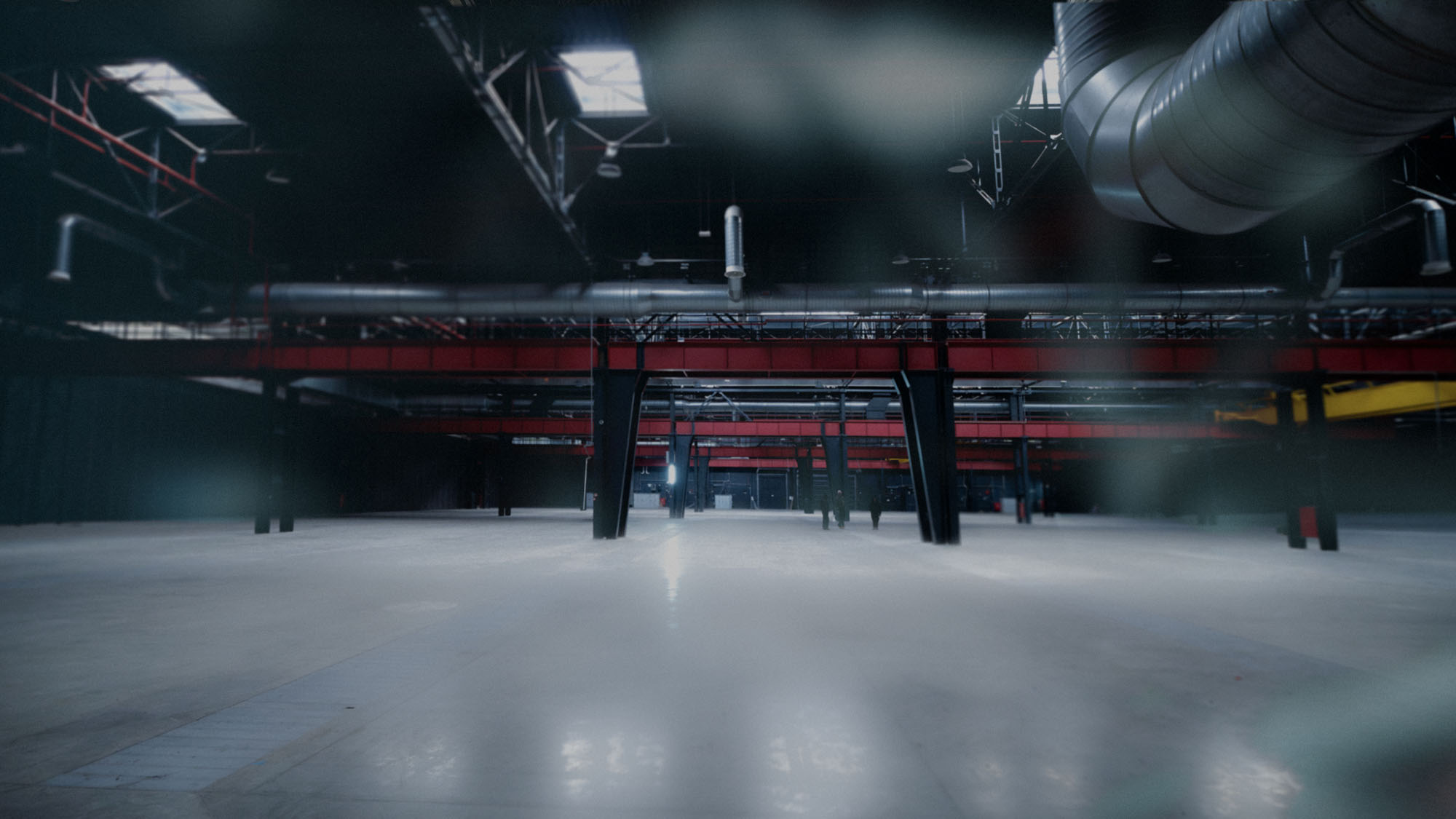 Wide empty industrial warehouse with red beams, metal pipes, and high ceilings.