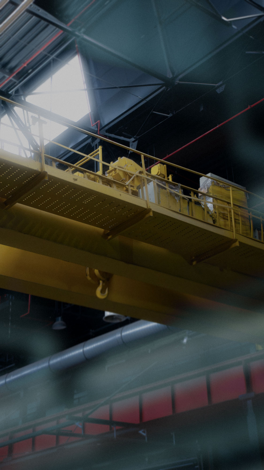 Yellow industrial crane with hook inside large factory building with metal beams and lighting.