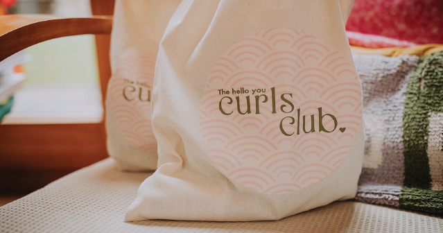 Two canvas bags with pink circular logos reading 'The hello you curls club' placed on a chair.