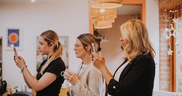 Three women looking at their phones in a warmly lit room with hanging lamps and decorative wall art.
