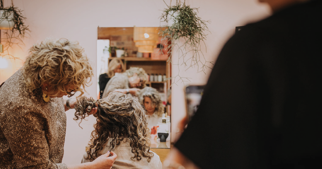 Hairdresser styling curly gray hair in a cozy salon with plants and warm lighting.