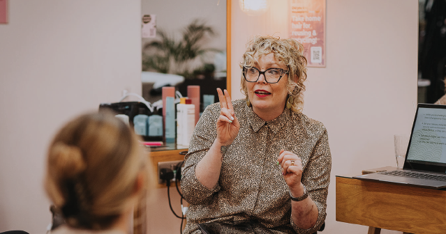 Curly-haired woman wearing glasses and patterned blouse speaking and gesturing with two fingers raised.