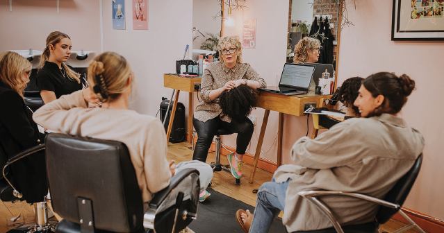 Group of people seated in a salon listening to a stylist holding a hairpiece during a consultation.