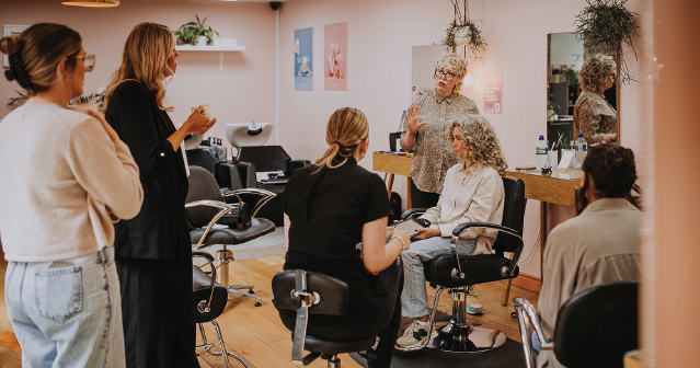 Hairstylist instructing a seated woman with curly hair in a salon while others watch attentively.