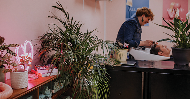 Woman washing a man's hair in a salon with plants and pink neon heart decor nearby.