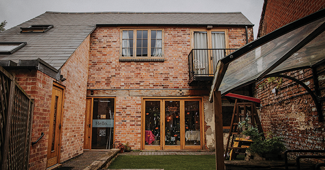 Brick two-story building with wooden doors, a small balcony, and a glass canopy over plants.