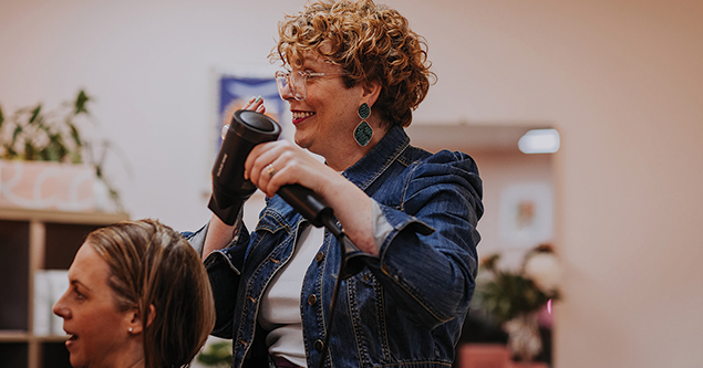 Smiling hairstylist with curly hair dries a seated client's hair using a black hairdryer.