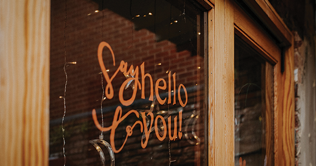 Window with orange lettering that says 'Say hello you!' reflected on a wooden frame and brick wall.