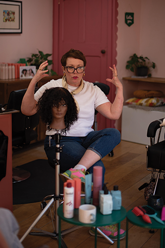 Woman with glasses demonstrating hair styling on a mannequin head in a salon setting.