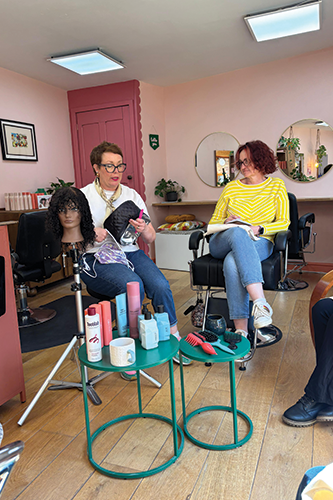 Two women seated in a salon with styling products and a mannequin head with curly hair nearby.