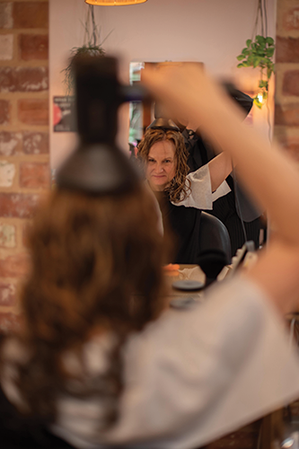 Woman drying curly hair with a blow dryer while looking into a salon mirror.