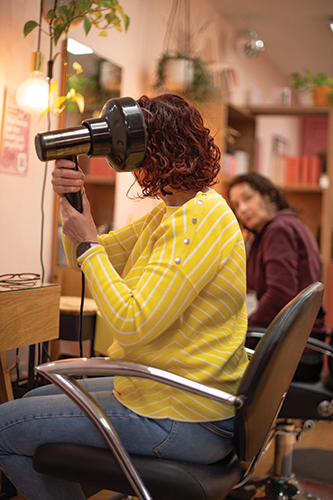 Person in yellow-striped sweater drying curly hair with a hairdryer in a salon chair.