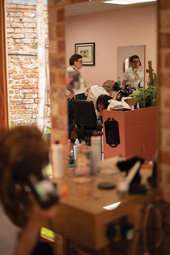 Hair salon interior with a stylist attending a client and another person looking in a mirror.